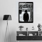 A black and white graphic poster featuring a hooded figure on a mountain path, with the text 'WOMEN'S SIGNATURE HOODIE' and 'IMPRINTED ICONOGRAPHY WEAR', displayed on a light gray wall above a modern white and dark wood console table with a black tripod floor lamp.
