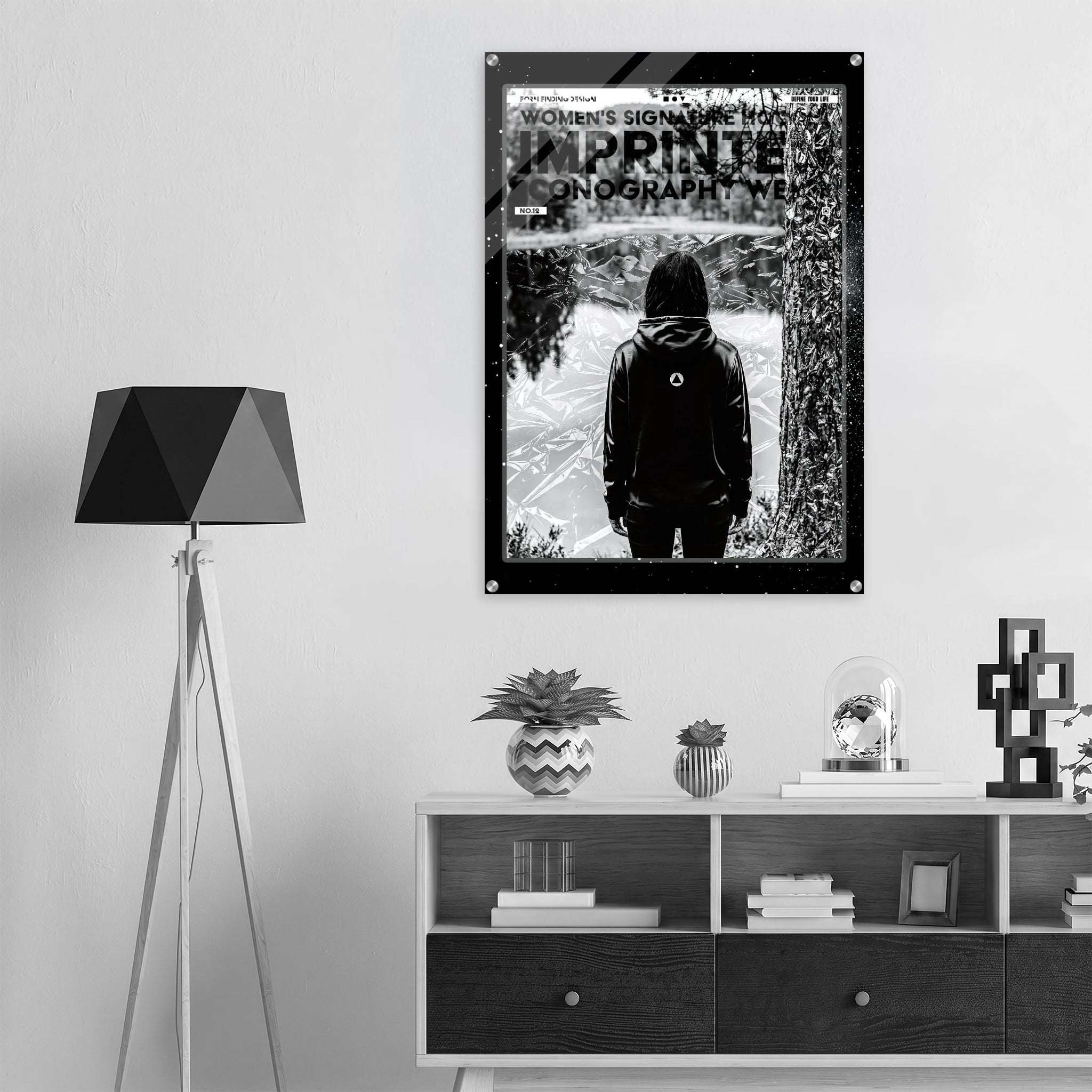 A black and white graphic poster featuring a hooded figure on a mountain path, with the text 'WOMEN'S SIGNATURE HOODIE' and 'IMPRINTED ICONOGRAPHY WEAR', displayed on a light gray wall above a modern white and dark wood console table with a black tripod floor lamp.