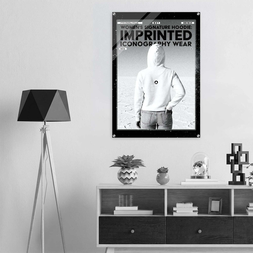 A black-framed poster featuring a black-and-white image of a person in a white hoodie with the text 'WOMEN'S SIGNATURE HOODIE IMPRINTED ICONOGRAPHY WEAR' displayed on a wall above a shelf in a modern room setting.