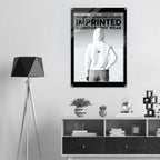 A black-framed poster featuring a black-and-white image of a person in a white hoodie with the text 'WOMEN'S SIGNATURE HOODIE IMPRINTED ICONOGRAPHY WEAR' displayed on a wall above a shelf in a modern room setting.