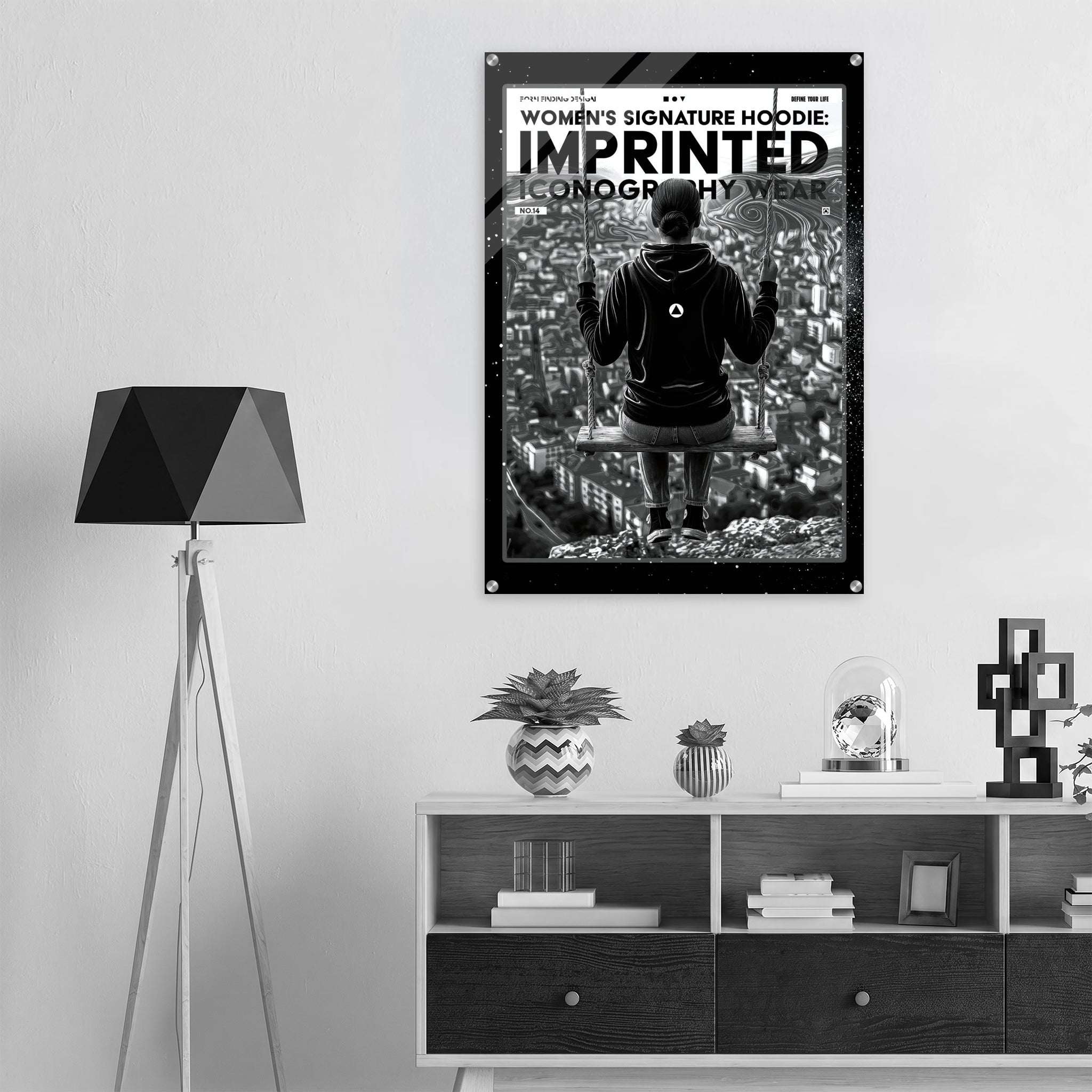 A black and white poster for the Women's Signature Hoodie 'Imprinted' is displayed on a light gray wall in a modern room setting above a wooden console table.