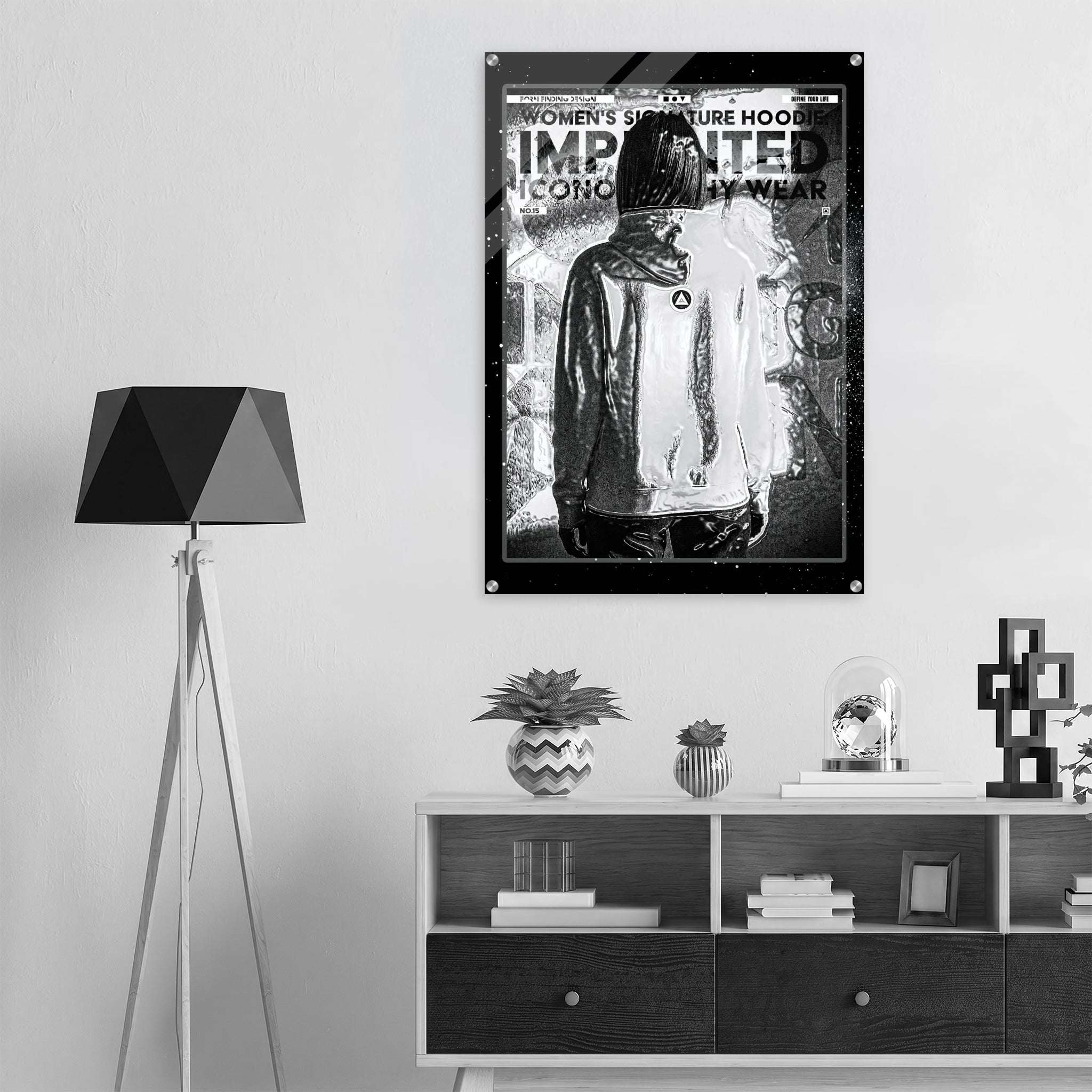 A black and white poster featuring a hooded figure with the text 'WOMEN'S SIGNATURE HOODIE IMPRINTED ICONOGRAPHY WEAR' displayed on a light gray wall above a shelf in a modern room setting.