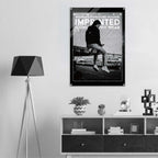 A black and white framed poster featuring a seated figure and the text 'WOMEN'S SIGNATURE HOODIE IMPRINTED ICONOGRAPHY WEAR' hanging on a light gray wall above a shelf in a modern interior setting.