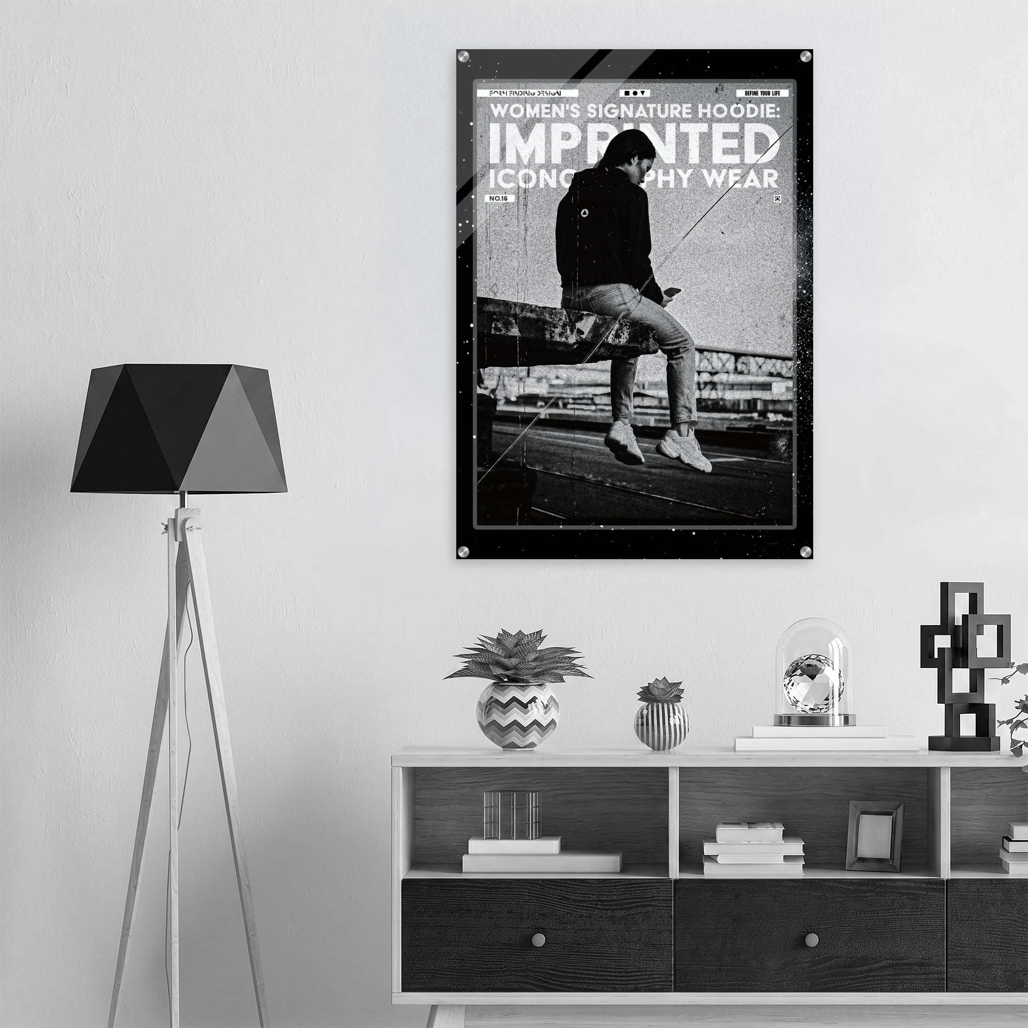 A black and white framed poster featuring a seated figure and the text 'WOMEN'S SIGNATURE HOODIE IMPRINTED ICONOGRAPHY WEAR' hanging on a light gray wall above a shelf in a modern interior setting.