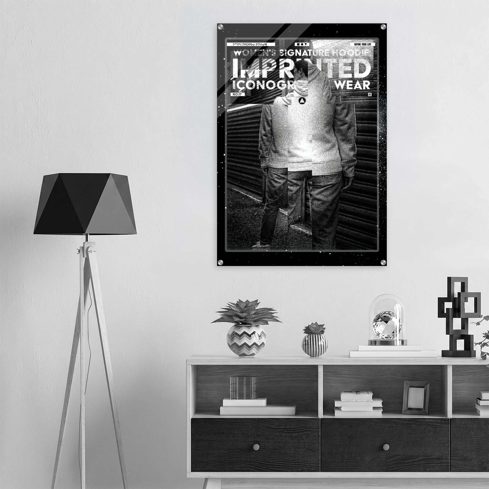 A black and white poster with the text 'WORKER'S SIGNATURE HOODIE: IMPRINTED ICONOGRAPHY WEAR' hangs on a light gray wall above a dark wood shelf in a modern room setting.