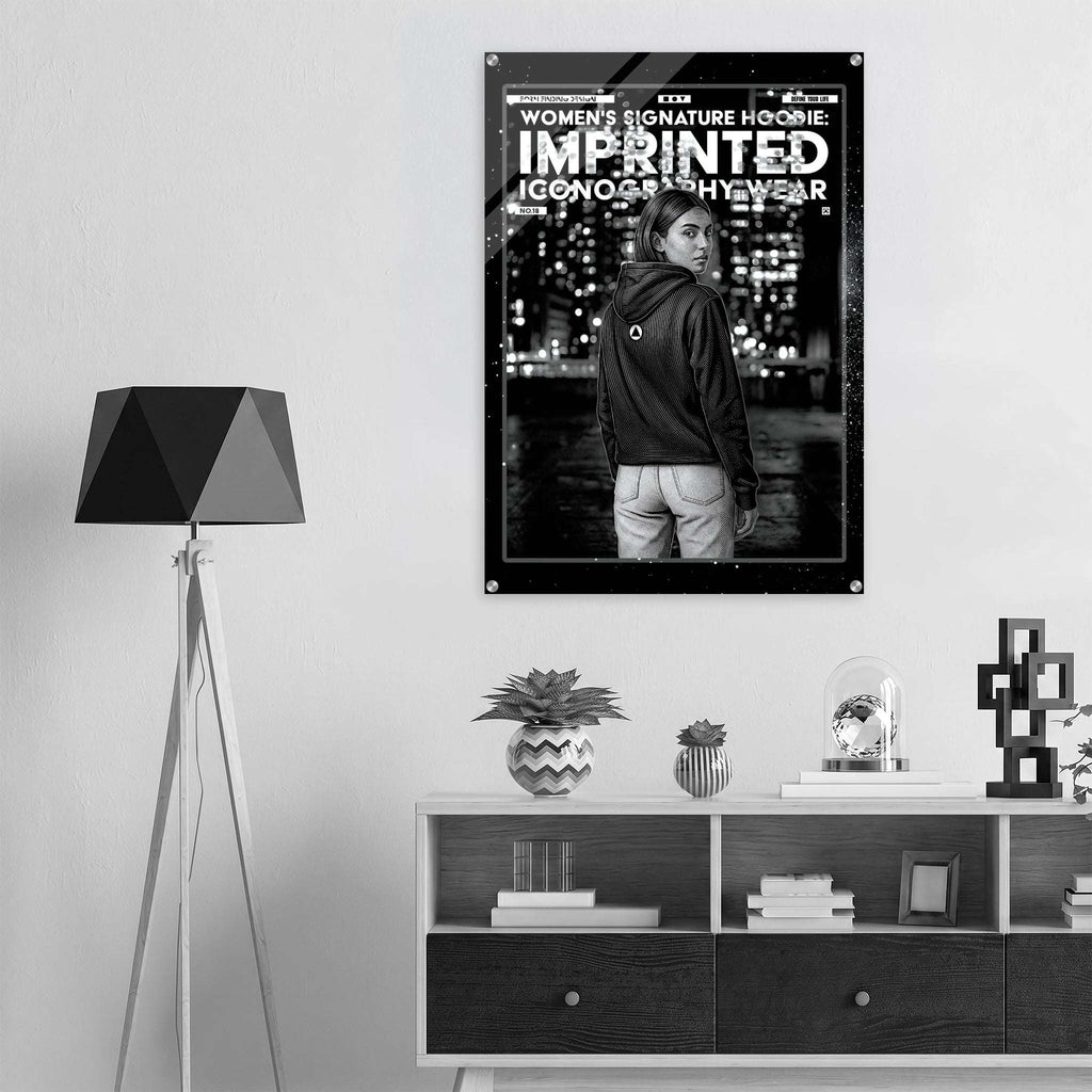 A black and white poster for the Women's Signature Hoodie 'Imprinted Iconography Wear' is displayed on a light gray wall in a modern room setting above a wooden console table.