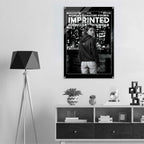A black and white poster for the Women's Signature Hoodie 'Imprinted Iconography Wear' is displayed on a light gray wall in a modern room setting above a wooden console table.