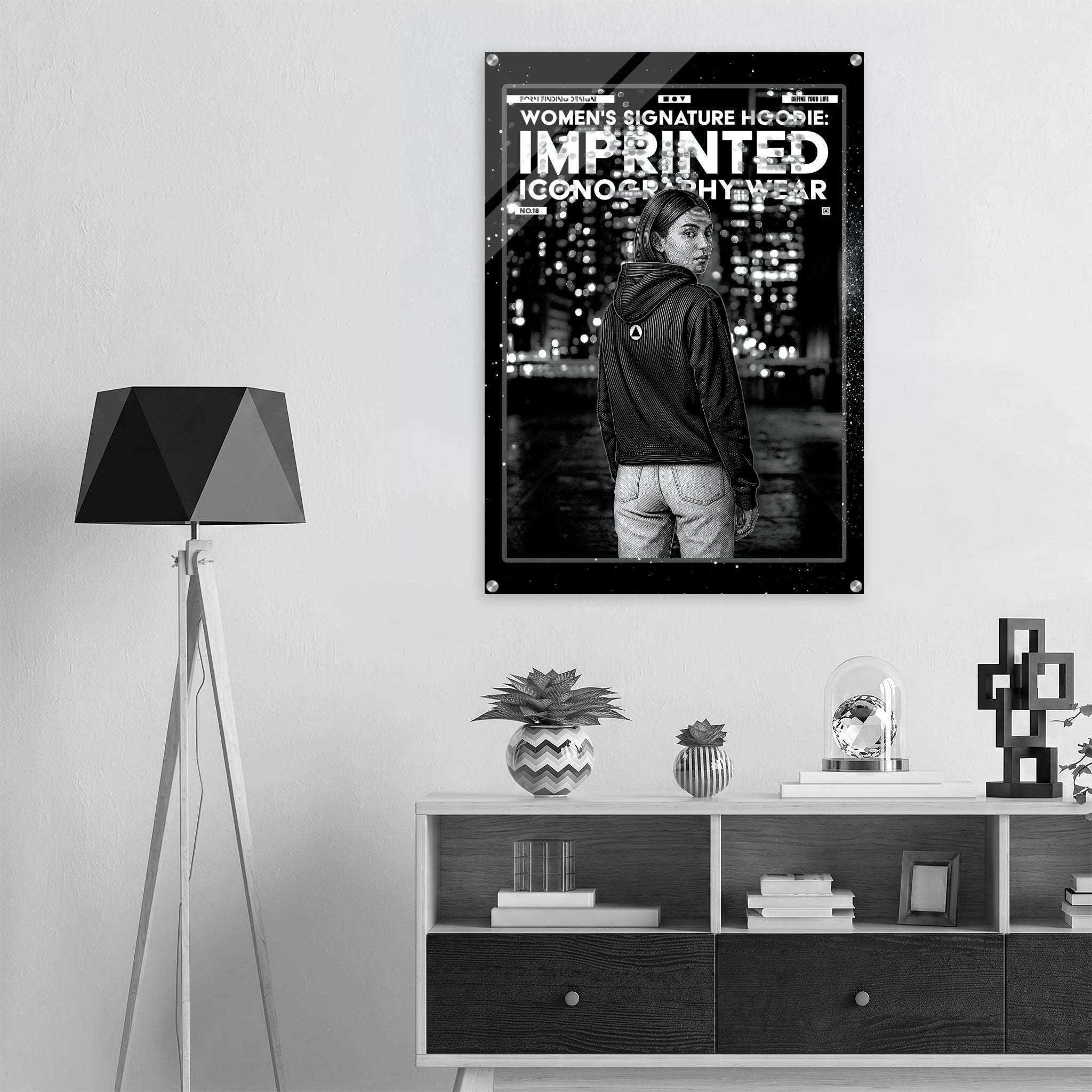 A black and white poster for the Women's Signature Hoodie 'Imprinted Iconography Wear' is displayed on a light gray wall in a modern room setting above a wooden console table.