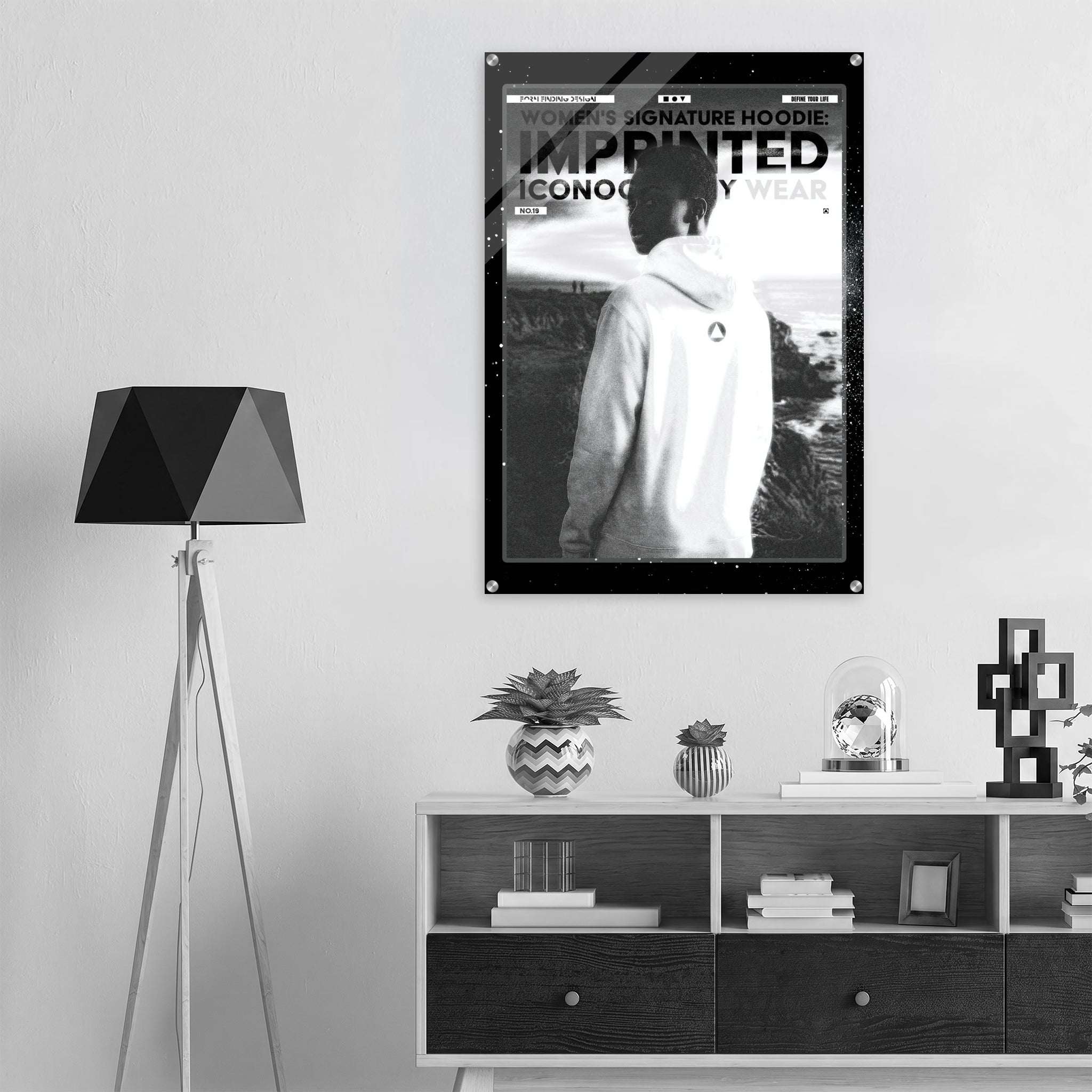 A black and white poster featuring a person in a hoodie hanging on a wall in a modern styled room with a tripod floor lamp and a low cabinet.