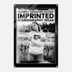 A black-framed art print or poster featuring a black-and-white photograph of a woman in a hoodie standing in a field, with bold text at the top reading 'WOMEN'S SIGNATURE HOODIE: IMPRINTED ICONOGRAPHY WEAR.'.