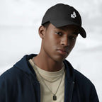 Male model wearing the black Under Armour x Form Fundamentals Stamp Cap (No.02) in a three‑quarter front pose; the cap’s curved brim and white Form Fundamentals logo are visible, styled with a navy hoodie over a beige top and layered chain in a neutral studio setting.