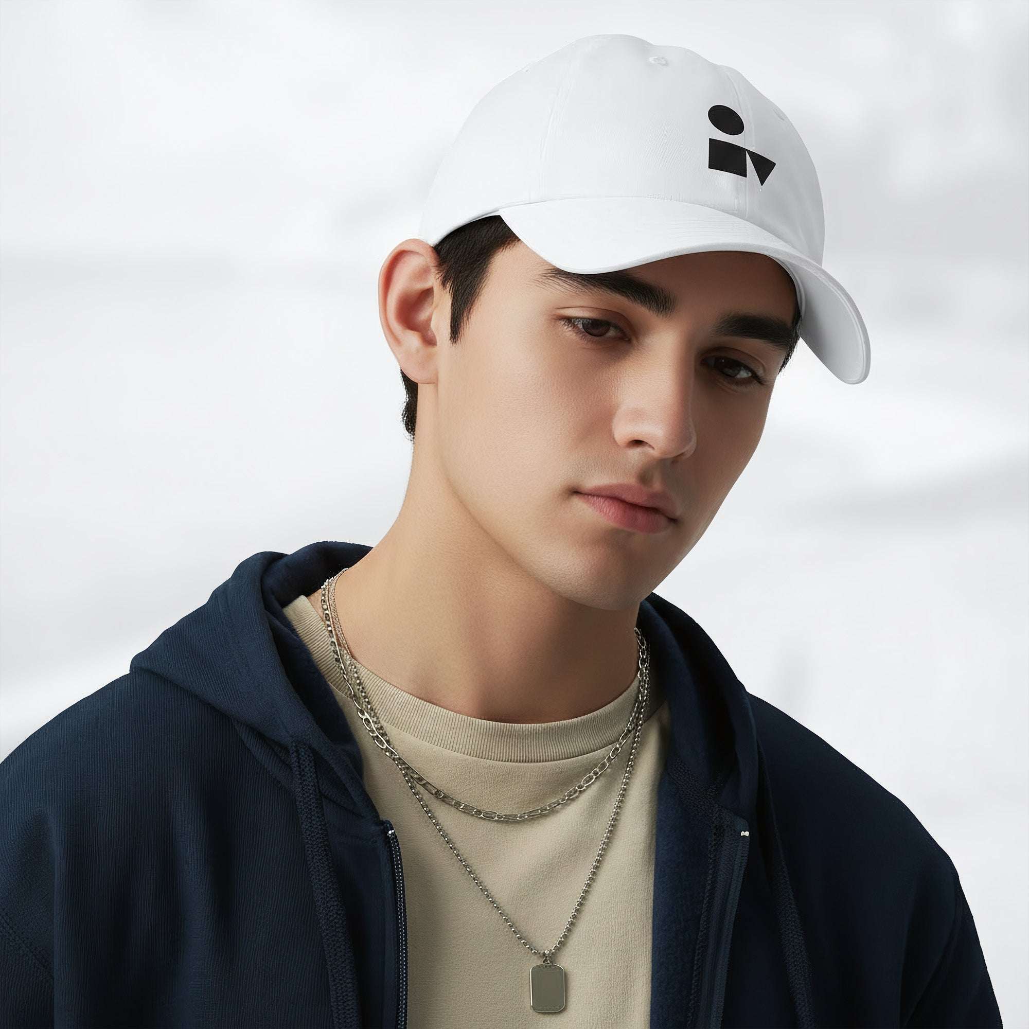 Male model wearing the white Under Armour x Form Fundamentals Stamp Cap (No.05) in a three‑quarter front pose; the cap’s curved brim and black Form Fundamentals logo are visible, styled with a navy hoodie over a beige top and layered chain in a neutral studio setting.