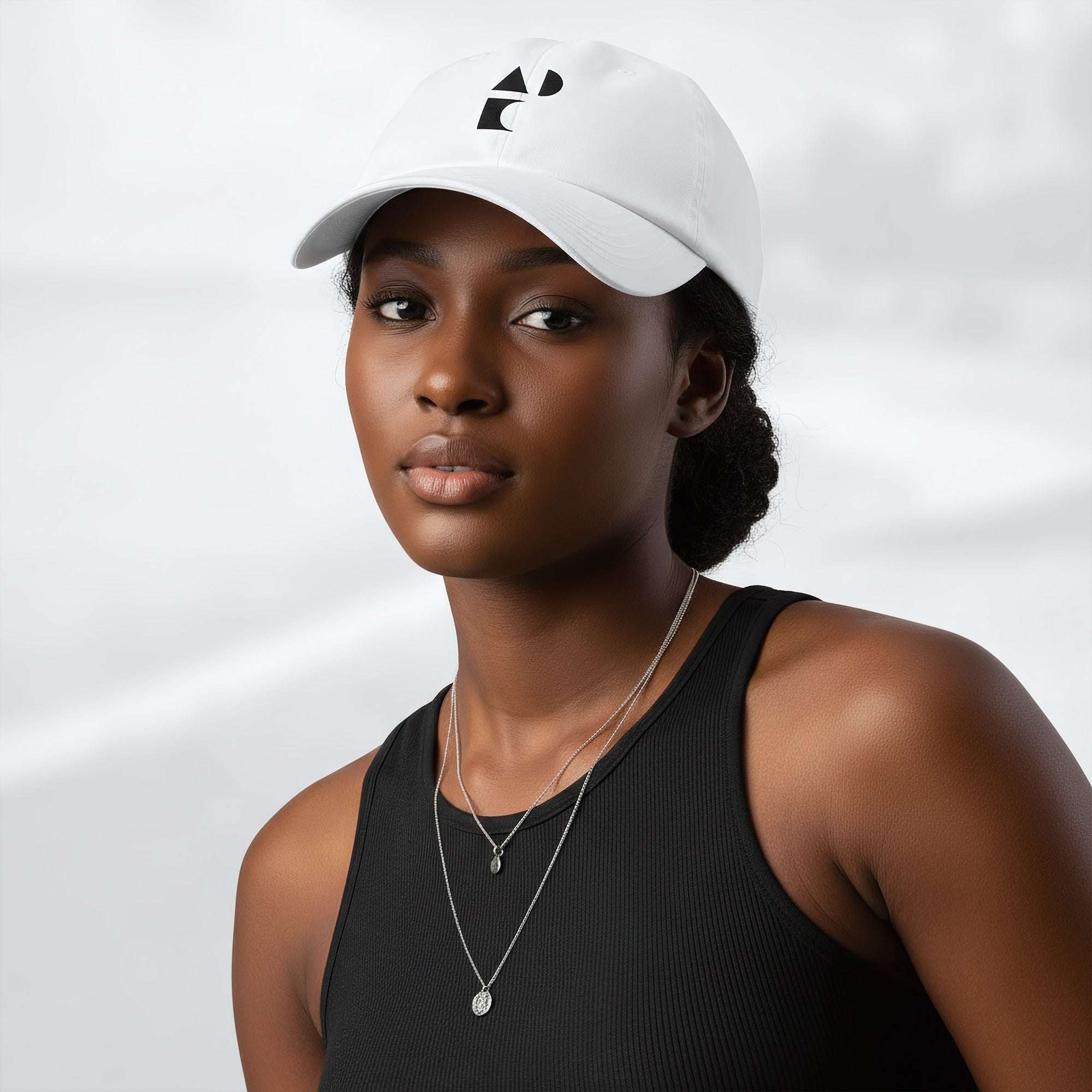 Female model wearing the white Under Armour x Form Fundamentals Stamp Cap (No.07); front view of the cap with black geometric emblem and curved brim, paired with a black tank top and layered necklaces against a softly blurred background.