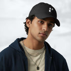 Male model wearing the black Under Armour x Form Fundamentals Stamp Cap (No.08) in a three‑quarter front pose; the cap’s curved brim and white Form Fundamentals logo are visible, styled with a navy hoodie over a beige top and layered chain in a neutral studio setting.