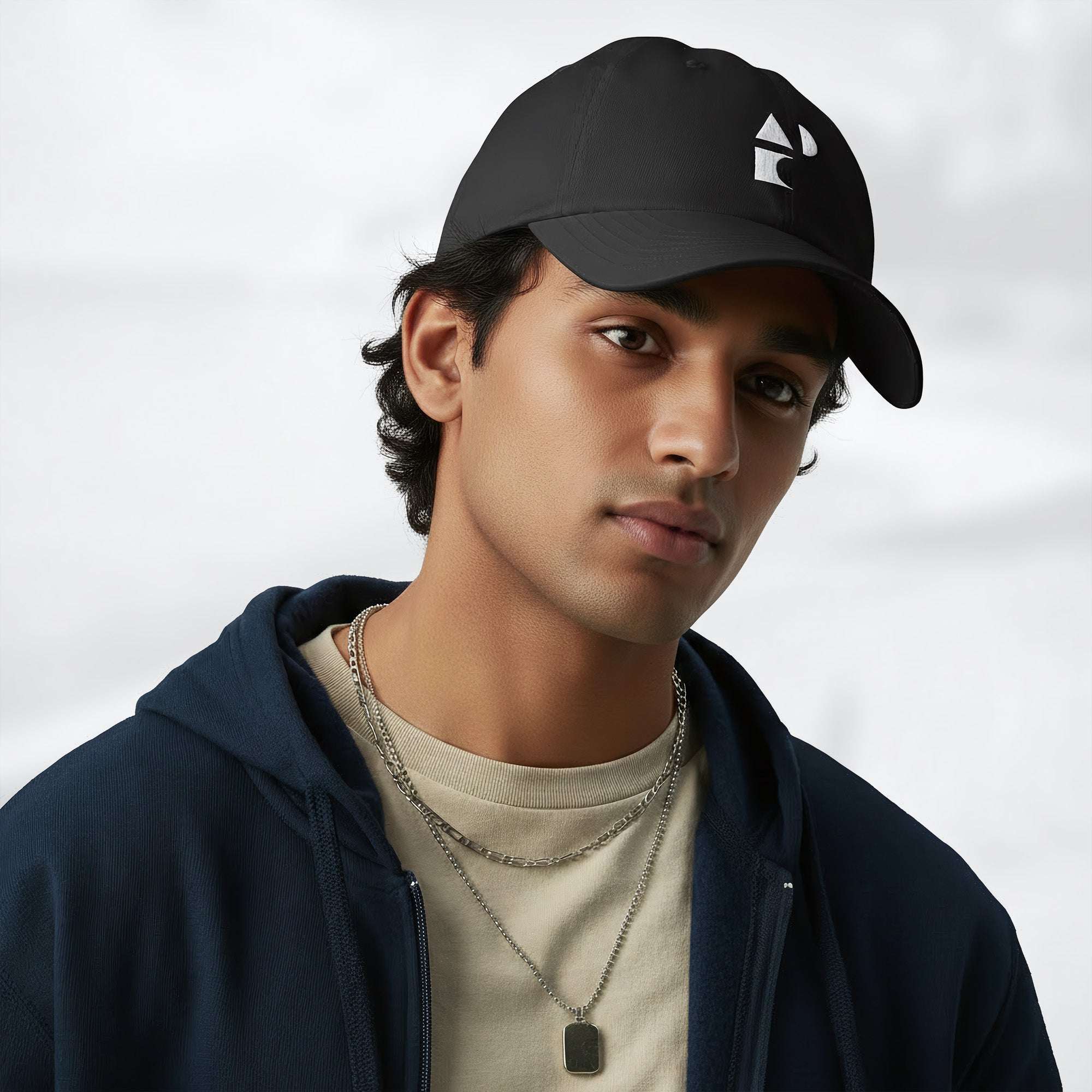 Male model wearing the black Under Armour x Form Fundamentals Stamp Cap (No.08) in a three‑quarter front pose; the cap’s curved brim and white Form Fundamentals logo are visible, styled with a navy hoodie over a beige top and layered chain in a neutral studio setting.