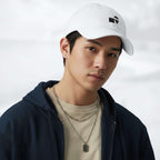 Male model wearing the white Under Armour x Form Fundamentals Stamp Cap (No.09) in a three‑quarter front pose; the cap’s curved brim and black Form Fundamentals logo are visible, styled with a navy hoodie over a beige top and layered chain in a neutral studio setting.