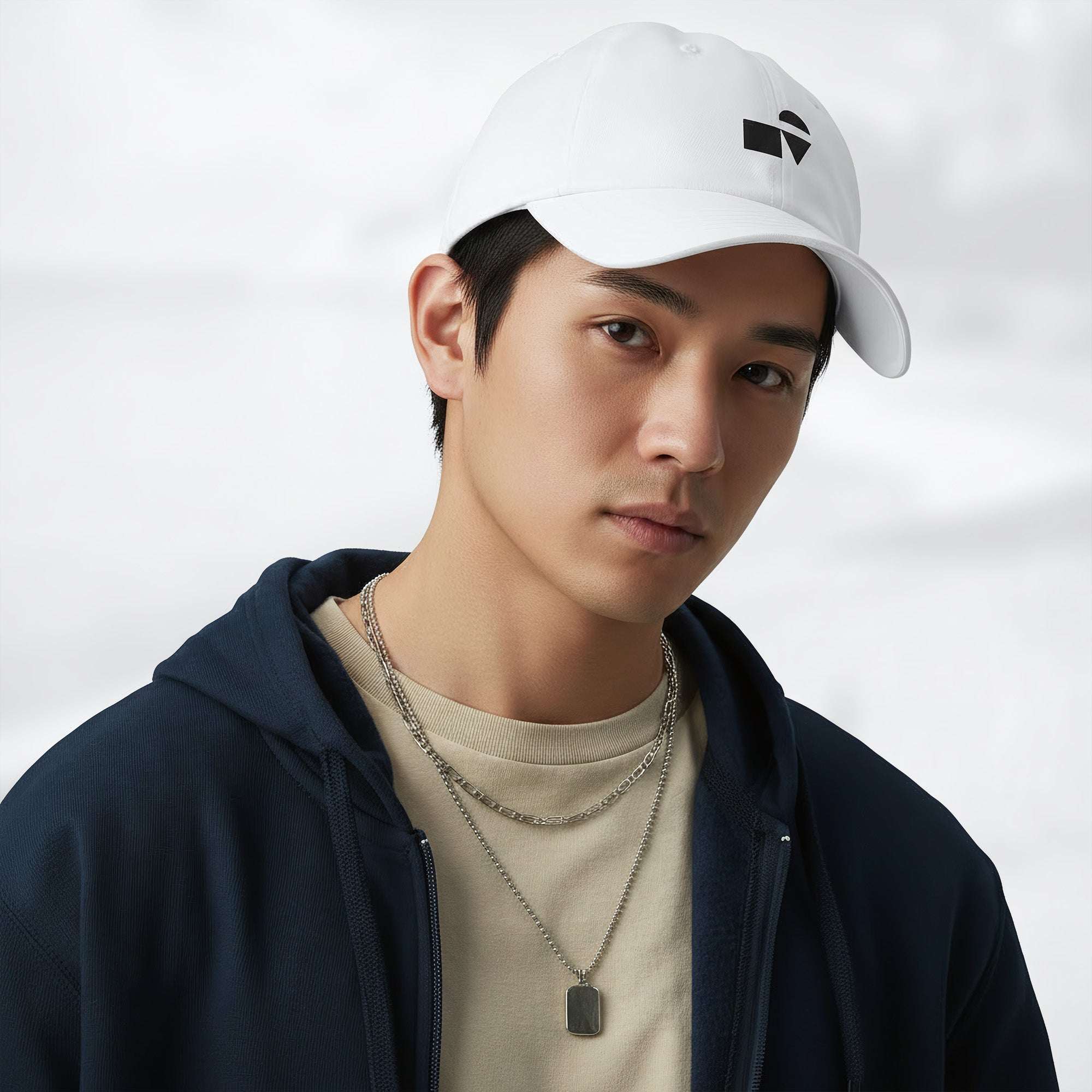 Male model wearing the white Under Armour x Form Fundamentals Stamp Cap (No.09) in a three‑quarter front pose; the cap’s curved brim and black Form Fundamentals logo are visible, styled with a navy hoodie over a beige top and layered chain in a neutral studio setting.