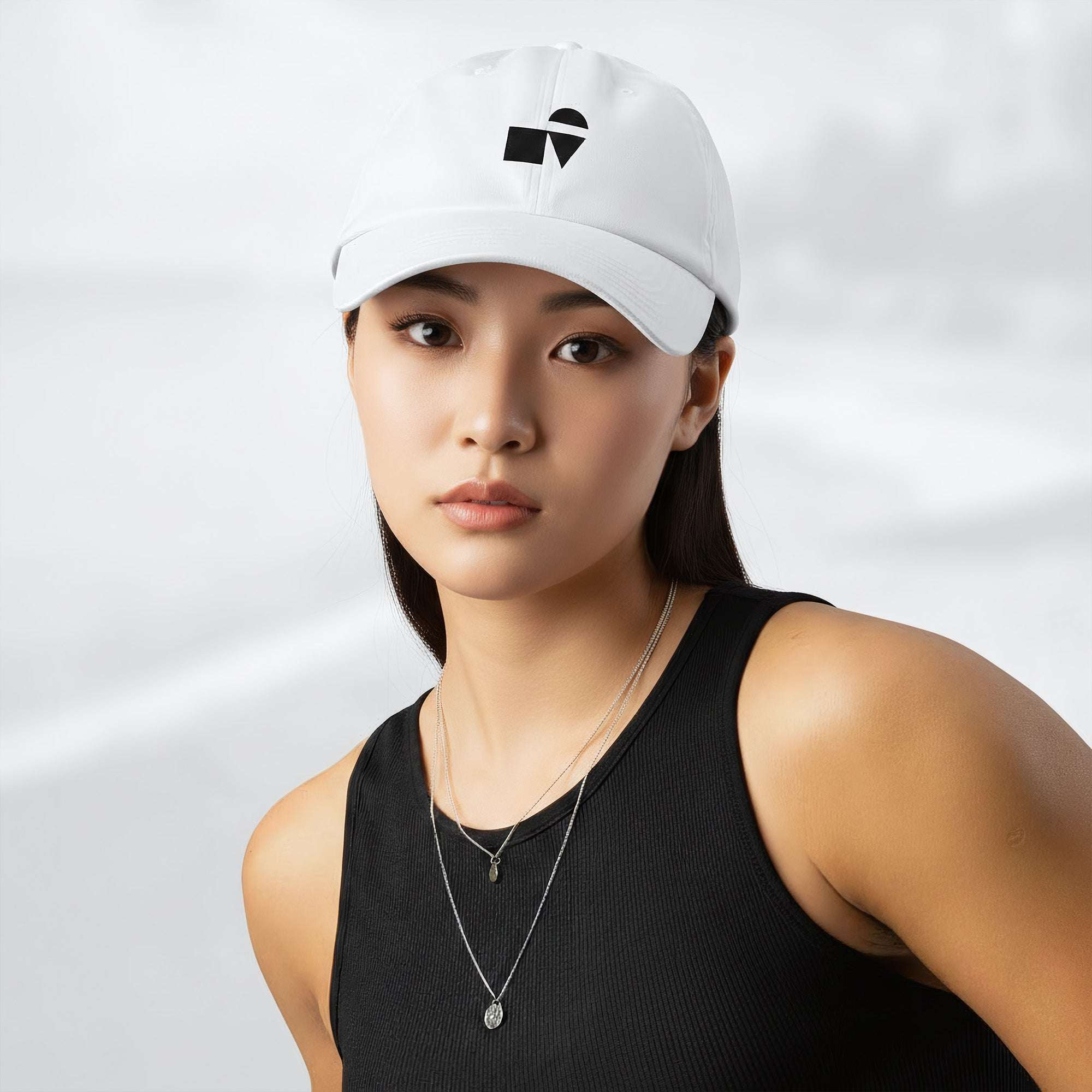 Female model wearing the white Under Armour x Form Fundamentals Stamp Cap (No.09); front view of the cap with black geometric emblem and curved brim, paired with a black tank top and layered necklaces against a softly blurred background.