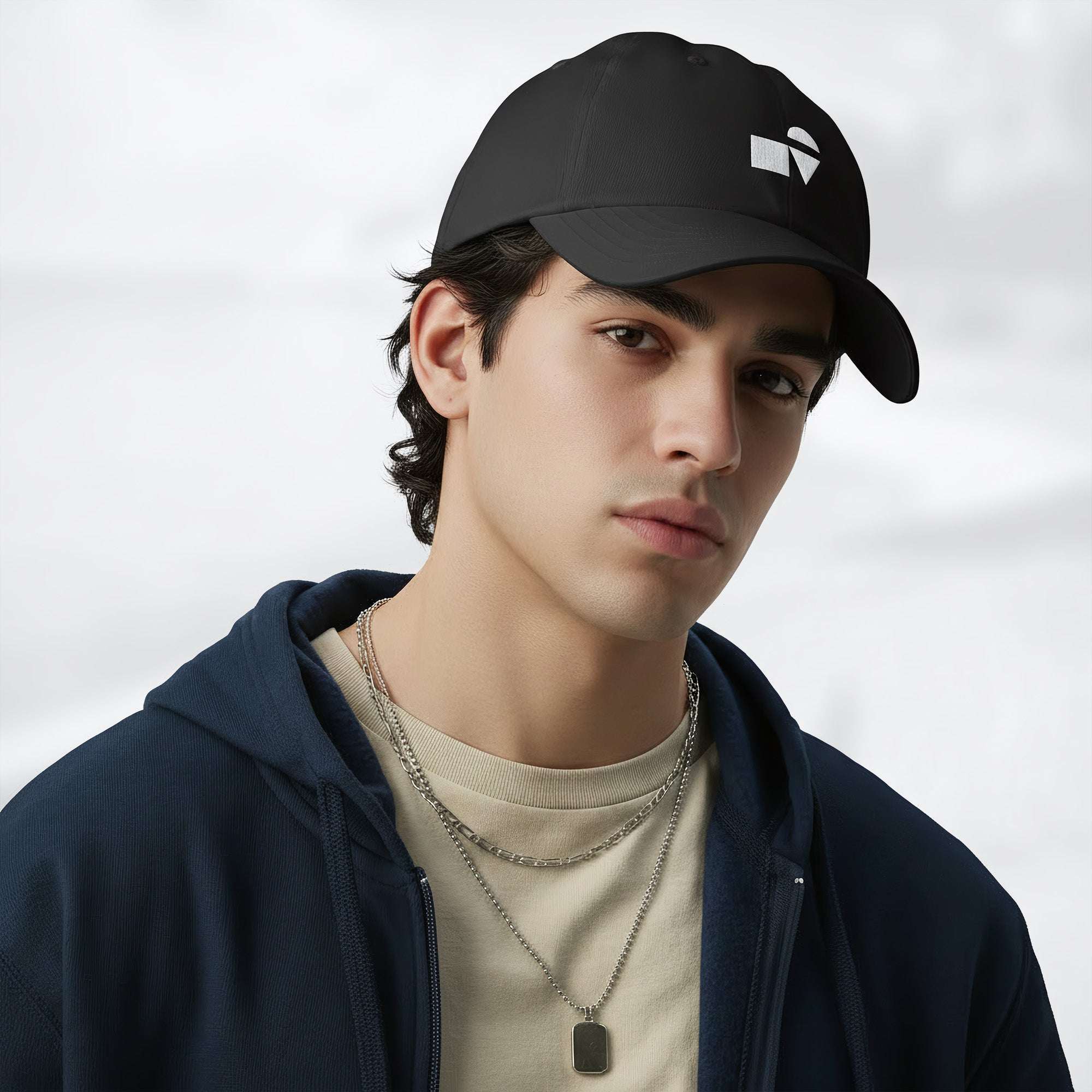 Male model wearing the black Under Armour x Form Fundamentals Stamp Cap (No.10) in a three‑quarter front pose; the cap’s curved brim and white Form Fundamentals logo are visible, styled with a navy hoodie over a beige top and layered chain in a neutral studio setting.