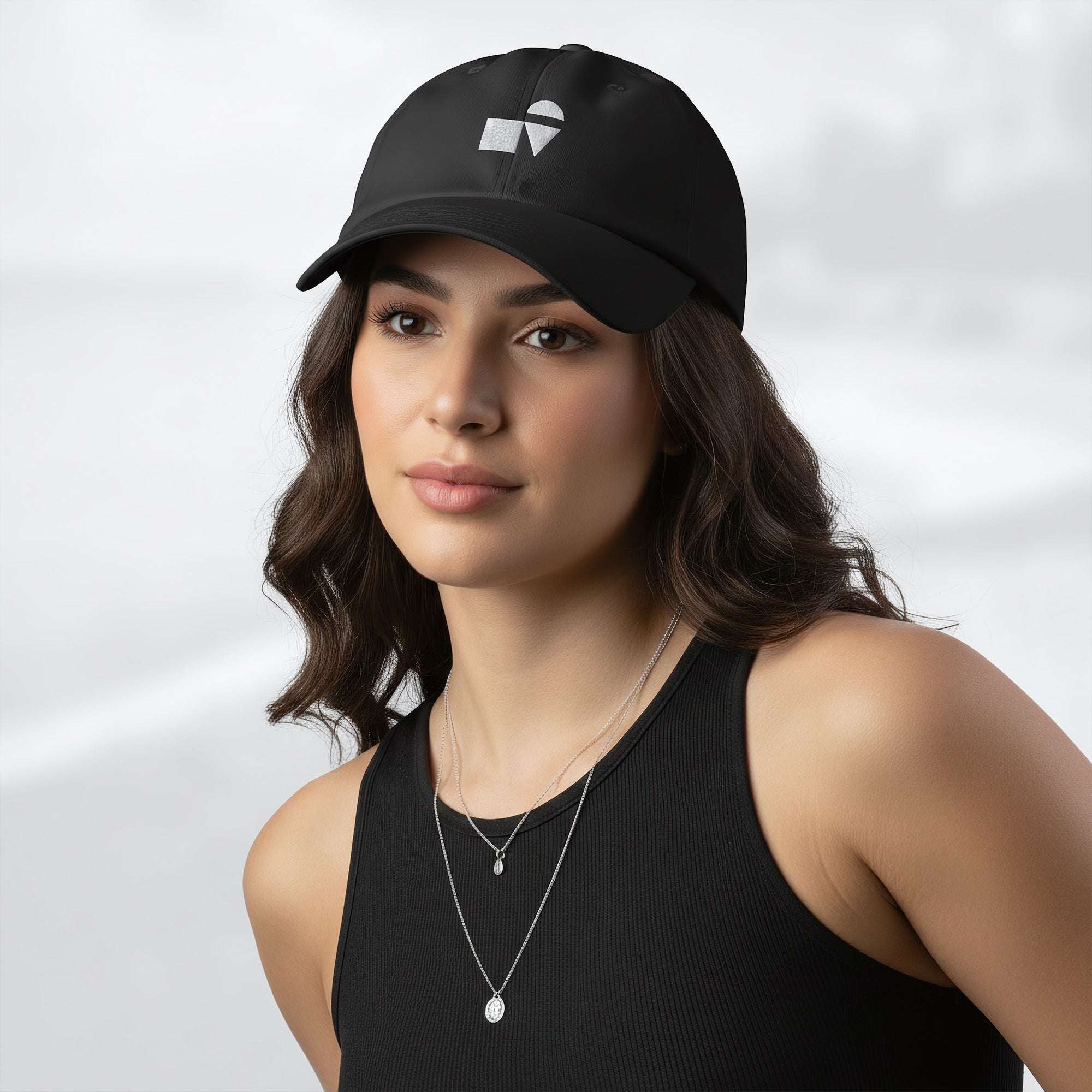 Female model wearing the black Under Armour x Form Fundamentals Stamp Cap (No.10); front view of the cap with white geometric emblem and curved brim, paired with a black tank top and layered necklaces against a softly blurred background.