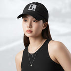 Female model wearing the black Under Armour x Form Fundamentals Stamp Cap (No.14); front view of the cap with white geometric emblem and curved brim, paired with a black tank top and layered necklaces against a softly blurred background.