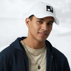 Male model wearing the white Under Armour x Form Fundamentals Stamp Cap (No.15) in a three‑quarter front pose; the cap’s curved brim and black Form Fundamentals logo are visible, styled with a navy hoodie over a beige top and layered chain in a neutral studio setting.