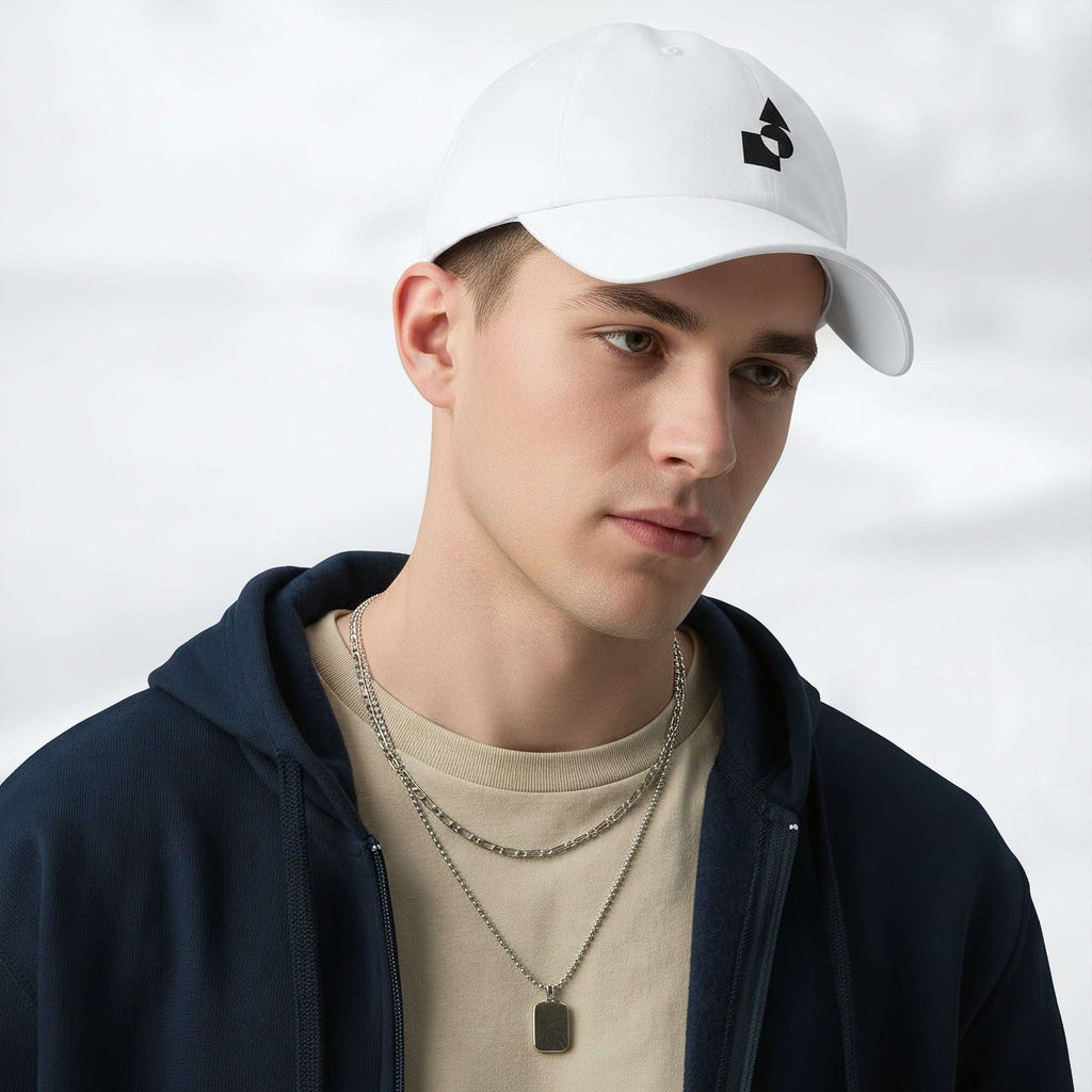 Male model wearing the white Under Armour x Form Fundamentals Stamp Cap in a three‑quarter front pose; the cap’s curved brim and black Form Fundamentals logo are visible, styled with a navy hoodie over a beige top and layered chain in a neutral studio setting.