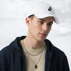 Male model wearing the white Under Armour x Form Fundamentals Stamp Cap in a three‑quarter front pose; the cap’s curved brim and black Form Fundamentals logo are visible, styled with a navy hoodie over a beige top and layered chain in a neutral studio setting.