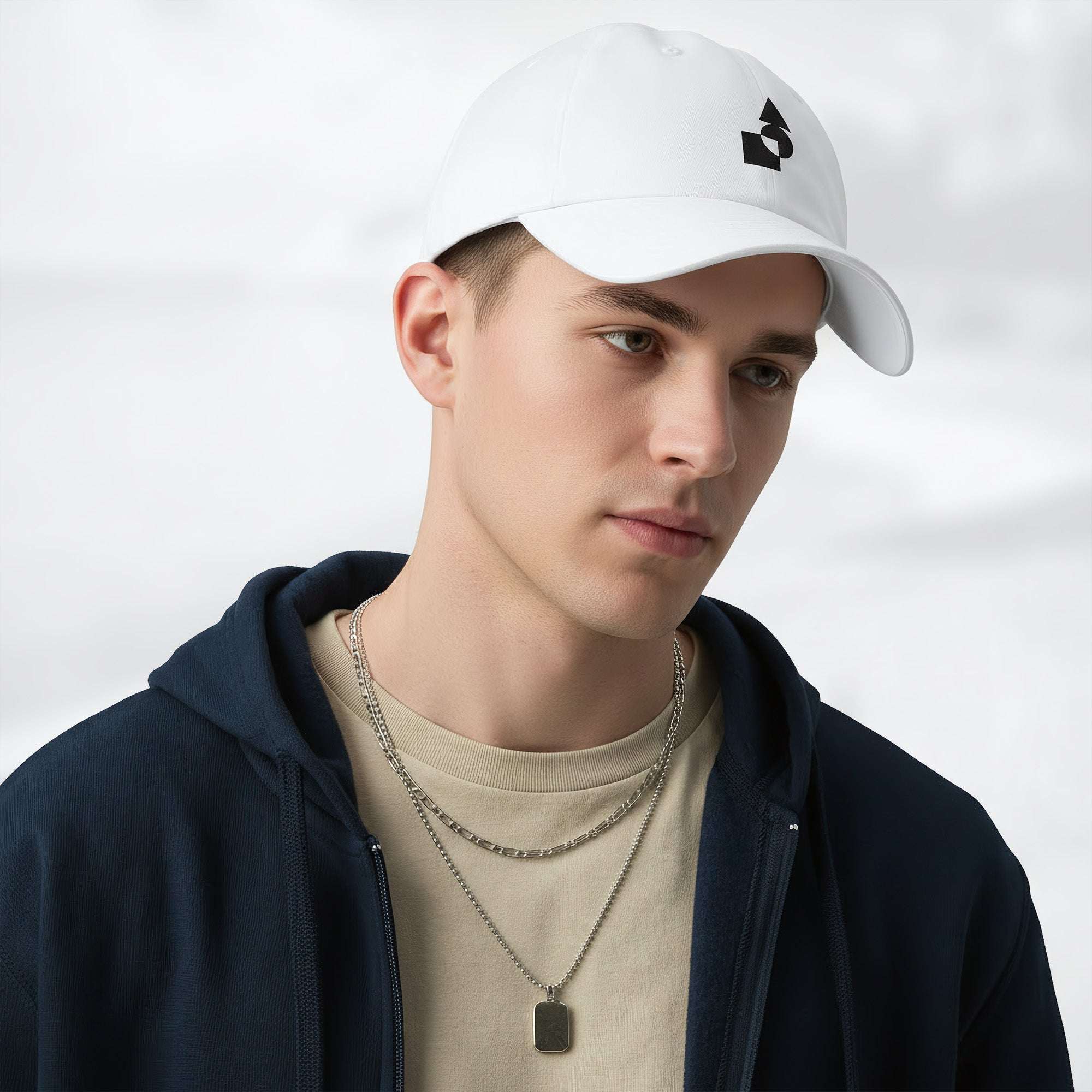 Male model wearing the white Under Armour x Form Fundamentals Stamp Cap in a three‑quarter front pose; the cap’s curved brim and black Form Fundamentals logo are visible, styled with a navy hoodie over a beige top and layered chain in a neutral studio setting.