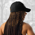 Rear‑side portrait of a woman with long braids wearing the black Under Armour x Title-3 Finding Logo Cap; the adjustable strap with a silver buckle is visible at the back while the curved bill faces left.