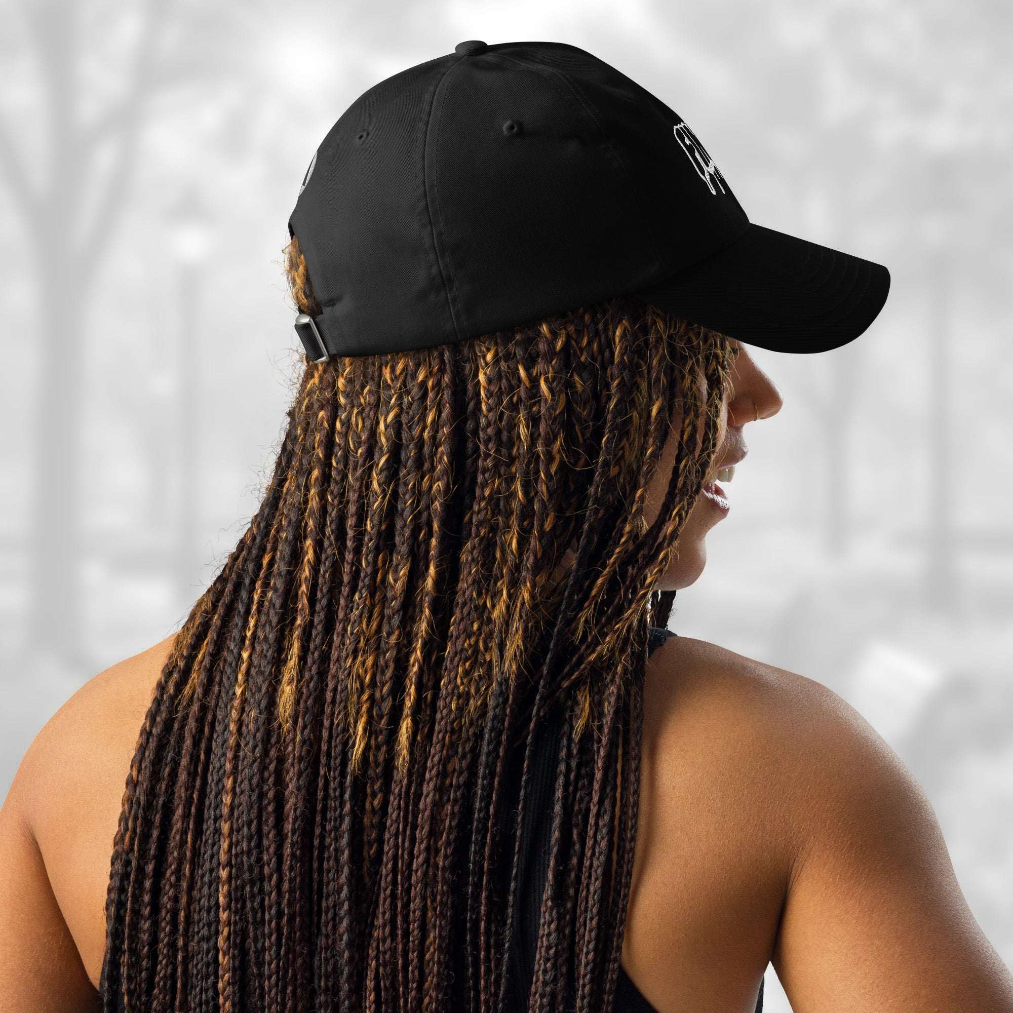 Rear‑side portrait of a woman with long braids wearing the black Under Armour x Title-3 Finding Logo Cap; the adjustable strap with a silver buckle is visible at the back while the curved bill faces left.