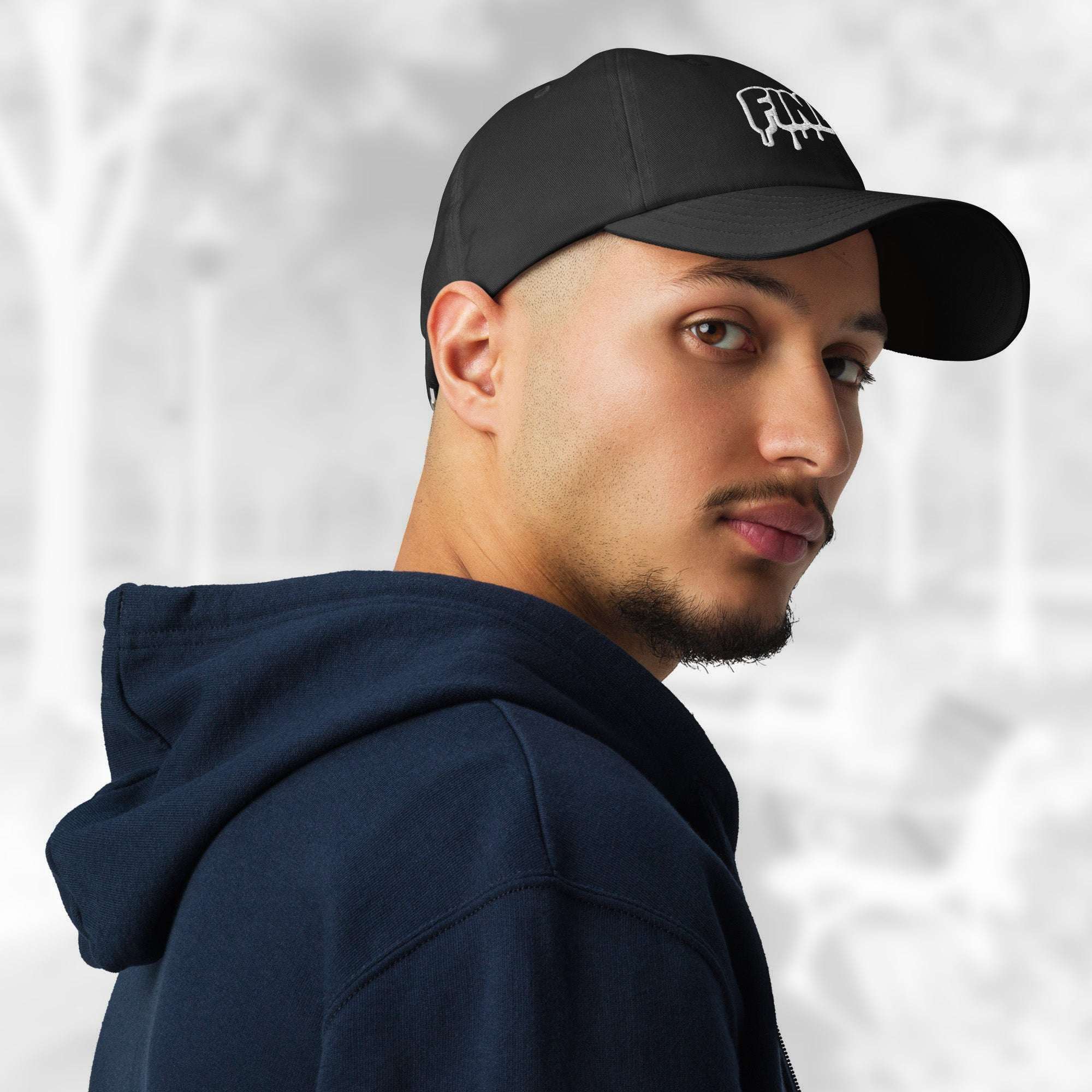 Three‑quarter portrait of a man wearing the Under Armour x Title-3 Finding Logo Cap—black cap with a partially visible white graffiti Finding graphic, curved visor; he wears a navy hoodie and layered chains against a softly blurred background.