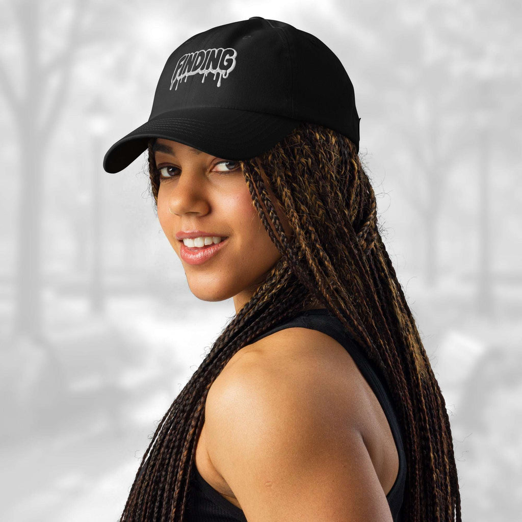 Three‑quarter front portrait of a woman wearing the Under Armour x Title-3 Finding Logo Cap—black cap with white graffiti‑style “Finding” emblem on the front, curved visor angled slightly down, her long braids draping over one shoulder.