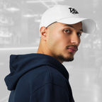 Three‑quarter portrait of a man wearing the Under Armour x Title-6 Design Logo Cap—white cap with a partially visible black graffiti Design graphic, curved visor; he wears a navy hoodie and layered chains against a softly blurred background.