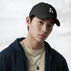 Male model wearing the black Under Armour x Form Fundamentals Stamp Cap (No.04) in a three‑quarter front pose; the cap’s curved brim and white Form Fundamentals logo are visible, styled with a navy hoodie over a beige top and layered chain in a neutral studio setting.