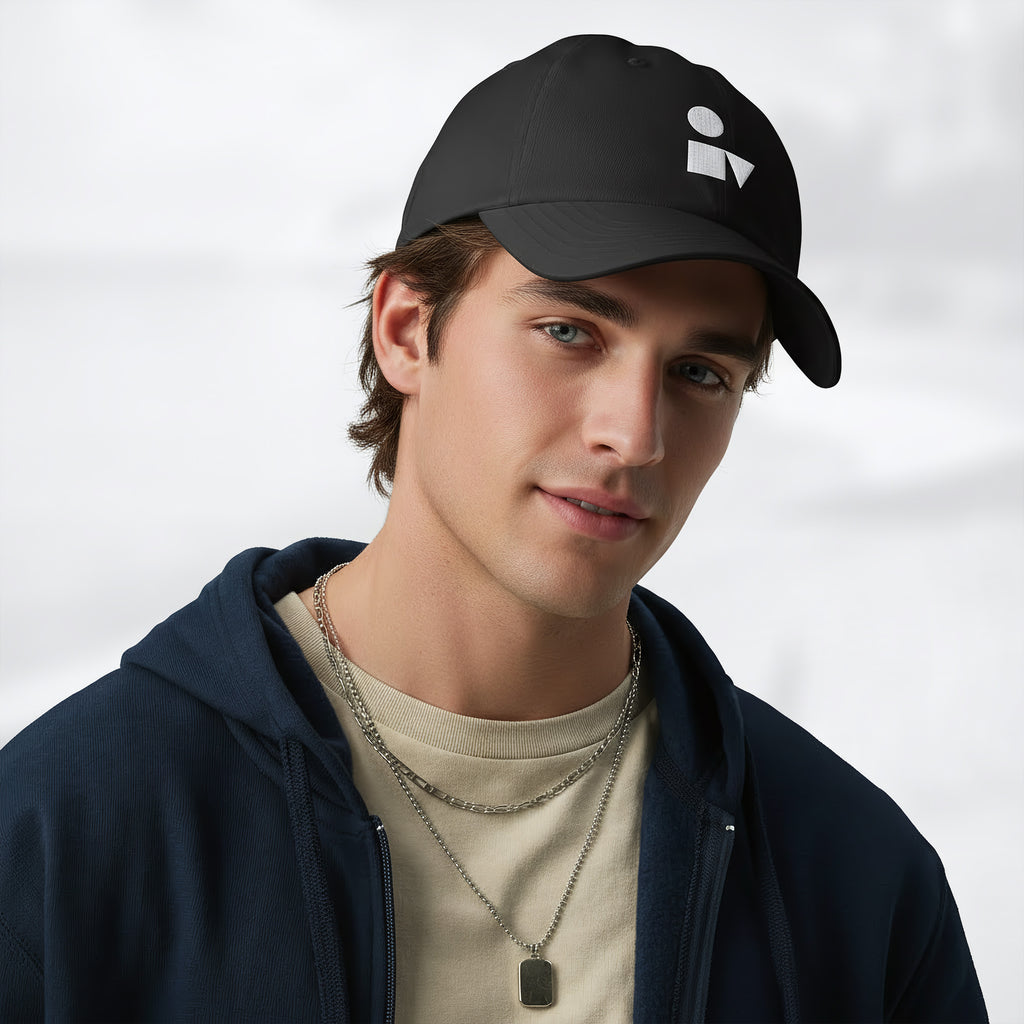 Male model wearing the black Under Armour x Form Fundamentals Stamp Cap (No.06) in a three‑quarter front pose; the cap’s curved brim and white Form Fundamentals logo are visible, styled with a navy hoodie over a beige top and layered chain in a neutral studio setting.