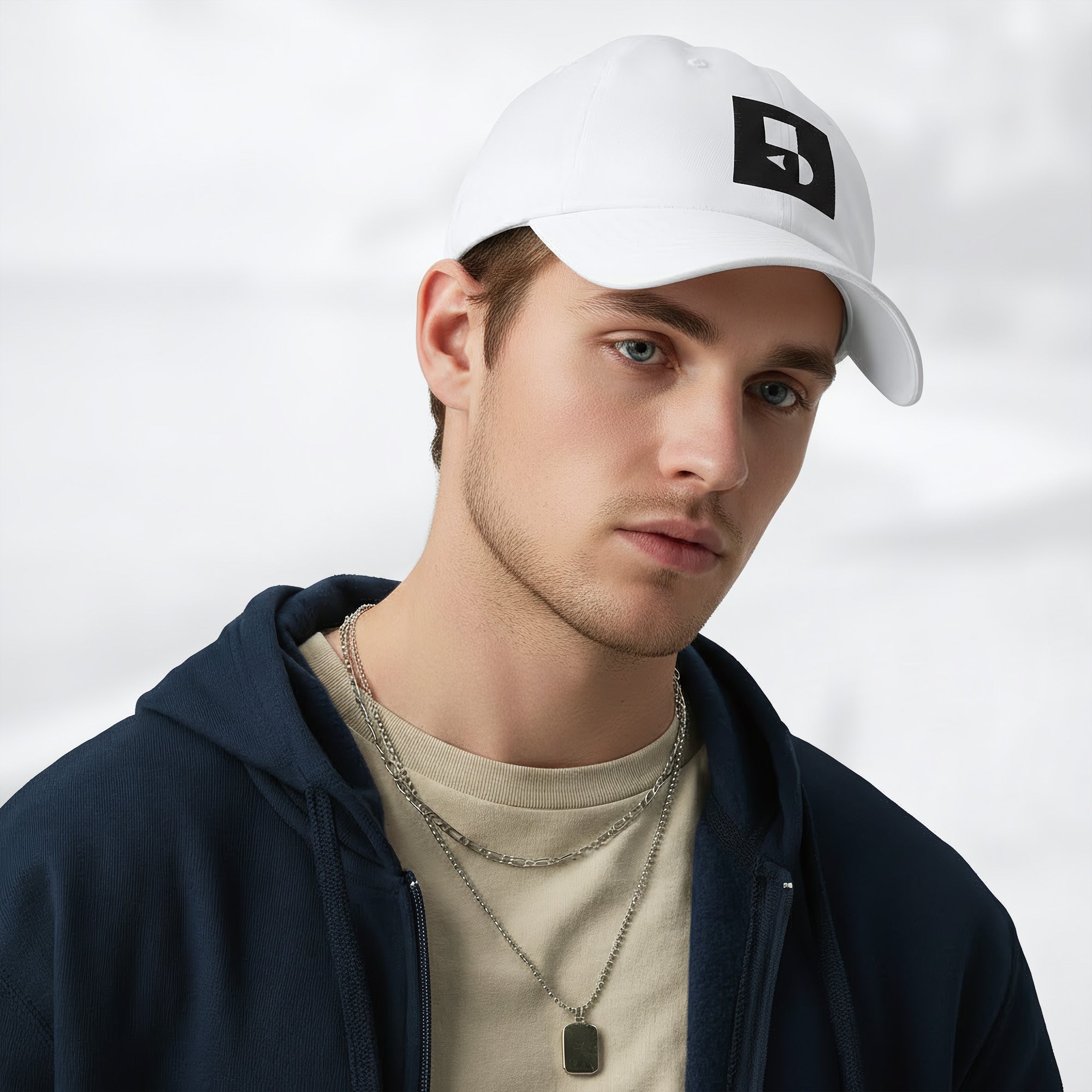 Male model wearing the white Under Armour x Form Fundamentals Stamp Cap (No.11) in a three‑quarter front pose; the cap’s curved brim and black Form Fundamentals logo are visible, styled with a navy hoodie over a beige top and layered chain in a neutral studio setting.