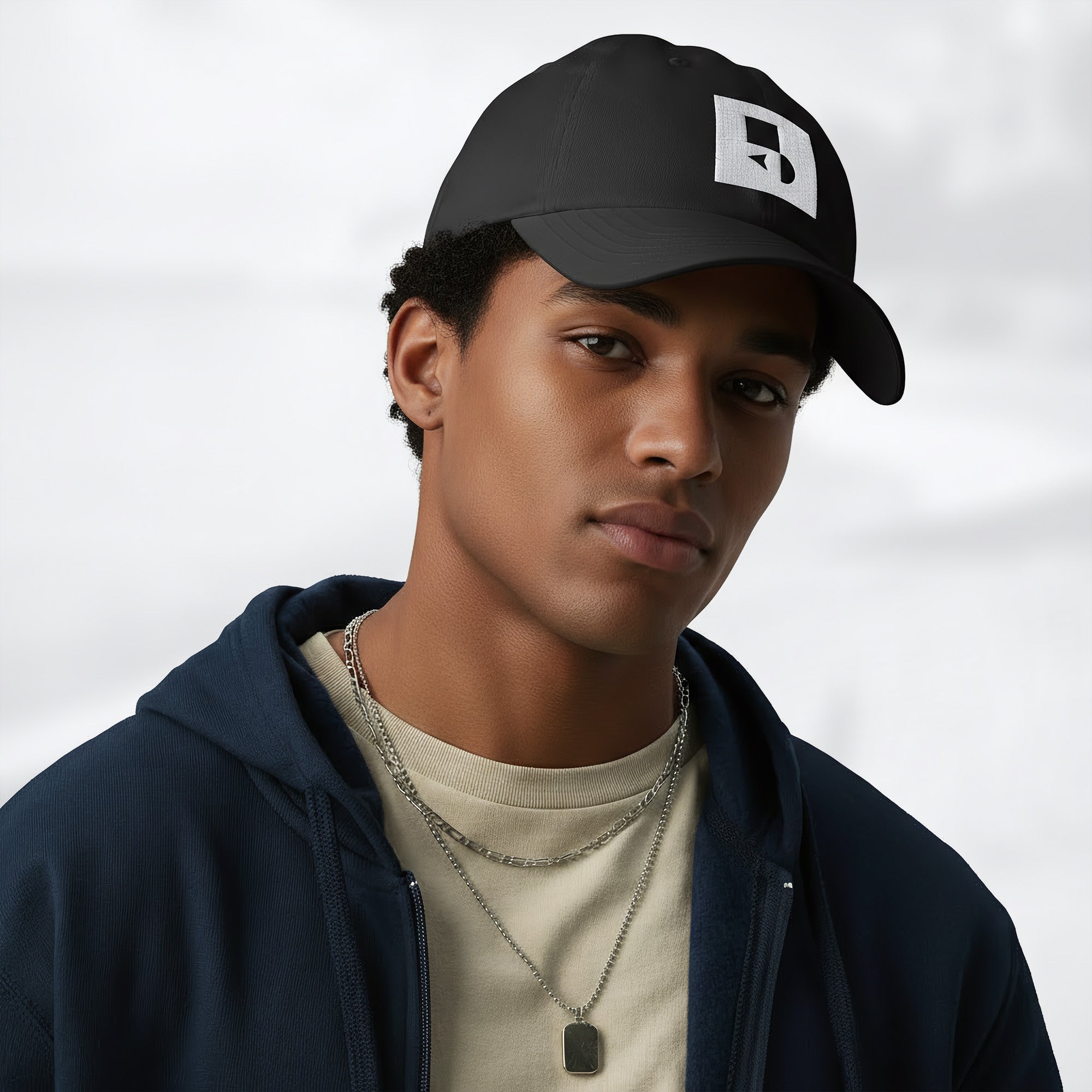 Male model wearing the black Under Armour x Form Fundamentals Stamp Cap (No.12) in a three‑quarter front pose; the cap’s curved brim and white Form Fundamentals logo are visible, styled with a navy hoodie over a beige top and layered chain in a neutral studio setting.