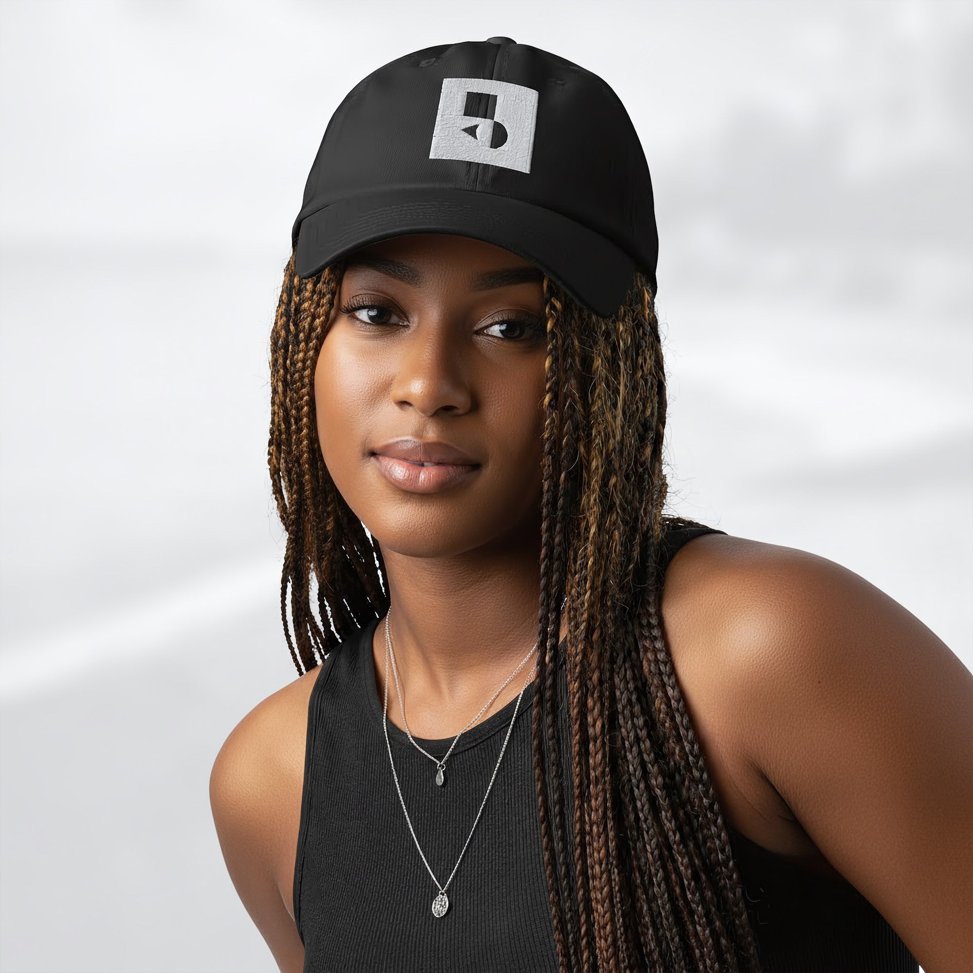 Female model wearing the black Under Armour x Form Fundamentals Stamp Cap (No.12); front view of the cap with white geometric emblem and curved brim, paired with a black tank top and layered necklaces against a softly blurred background.