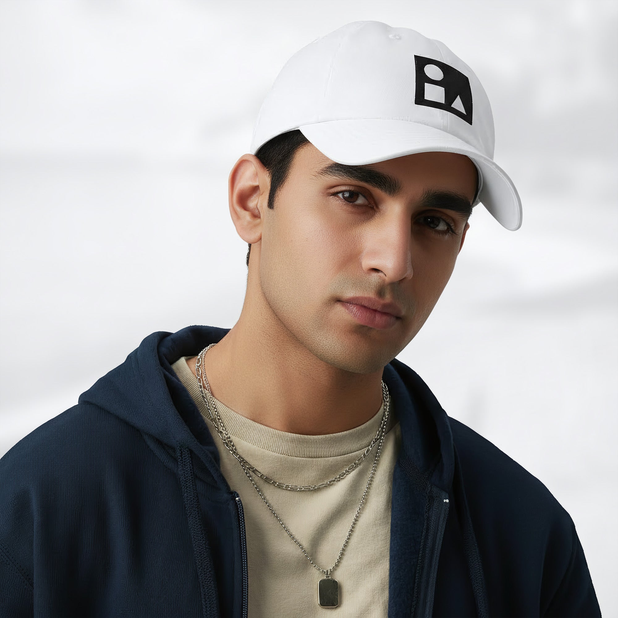Male model wearing the white Under Armour x Form Fundamentals Stamp Cap (No.13) in a three‑quarter front pose; the cap’s curved brim and black Form Fundamentals logo are visible, styled with a navy hoodie over a beige top and layered chain in a neutral studio setting.