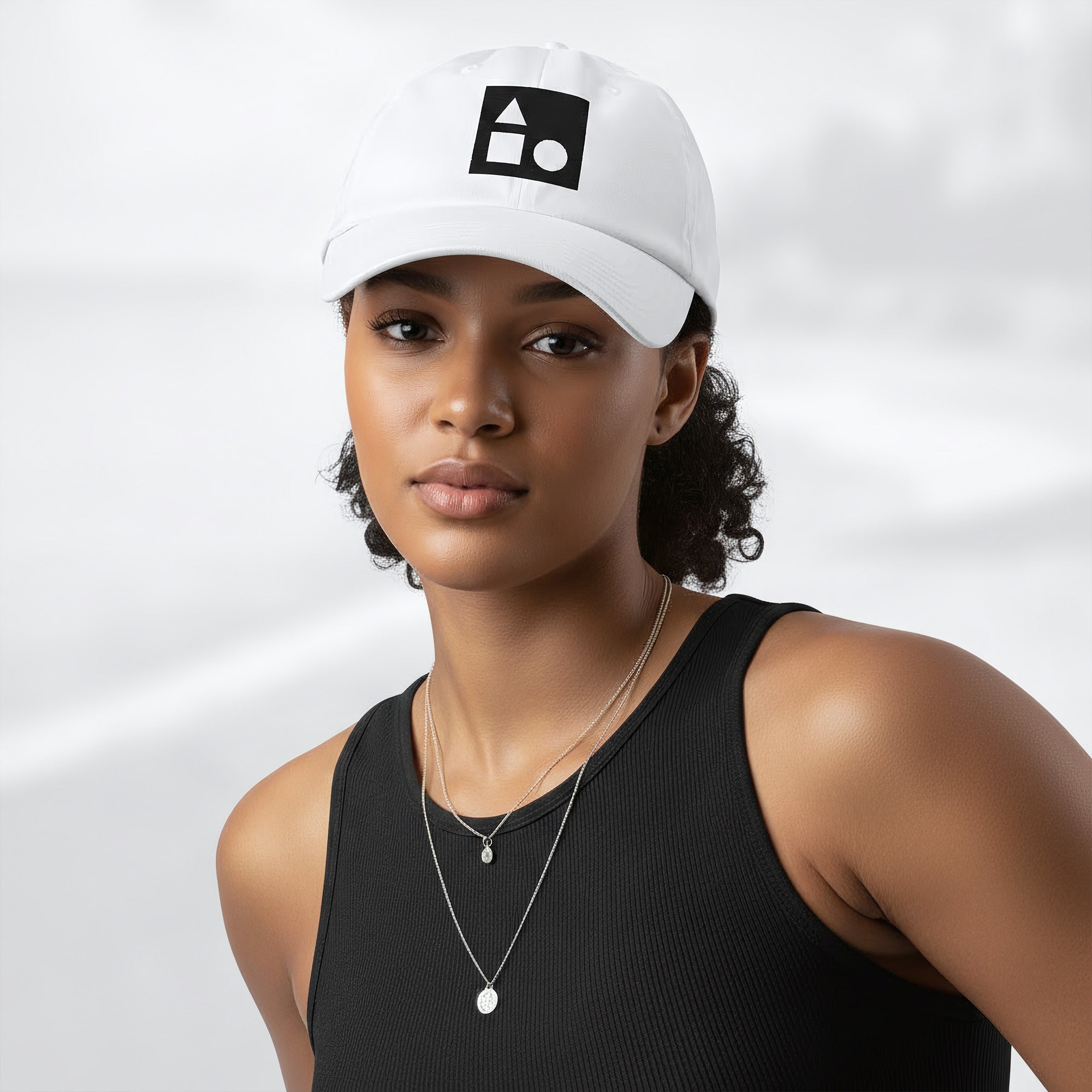 Female model wearing the white Under Armour x Form Fundamentals Stamp Cap (No.17); front view of the cap with black geometric emblem and curved brim, paired with a black tank top and layered necklaces against a softly blurred background.