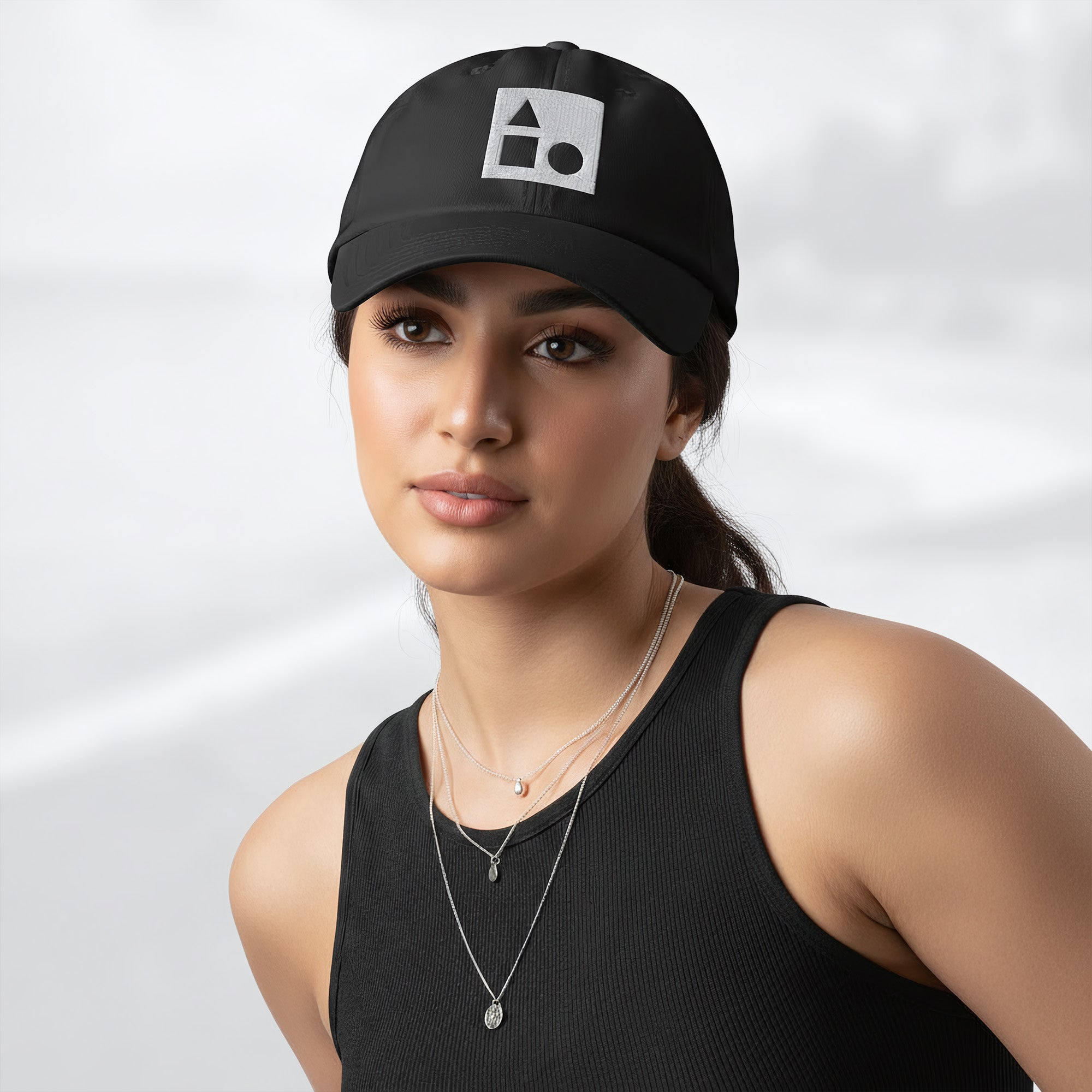 Female model wearing the black Under Armour x Form Fundamentals Stamp Cap (No.18); front view of the cap with white geometric emblem and curved brim, paired with a black tank top and layered necklaces against a softly blurred background.