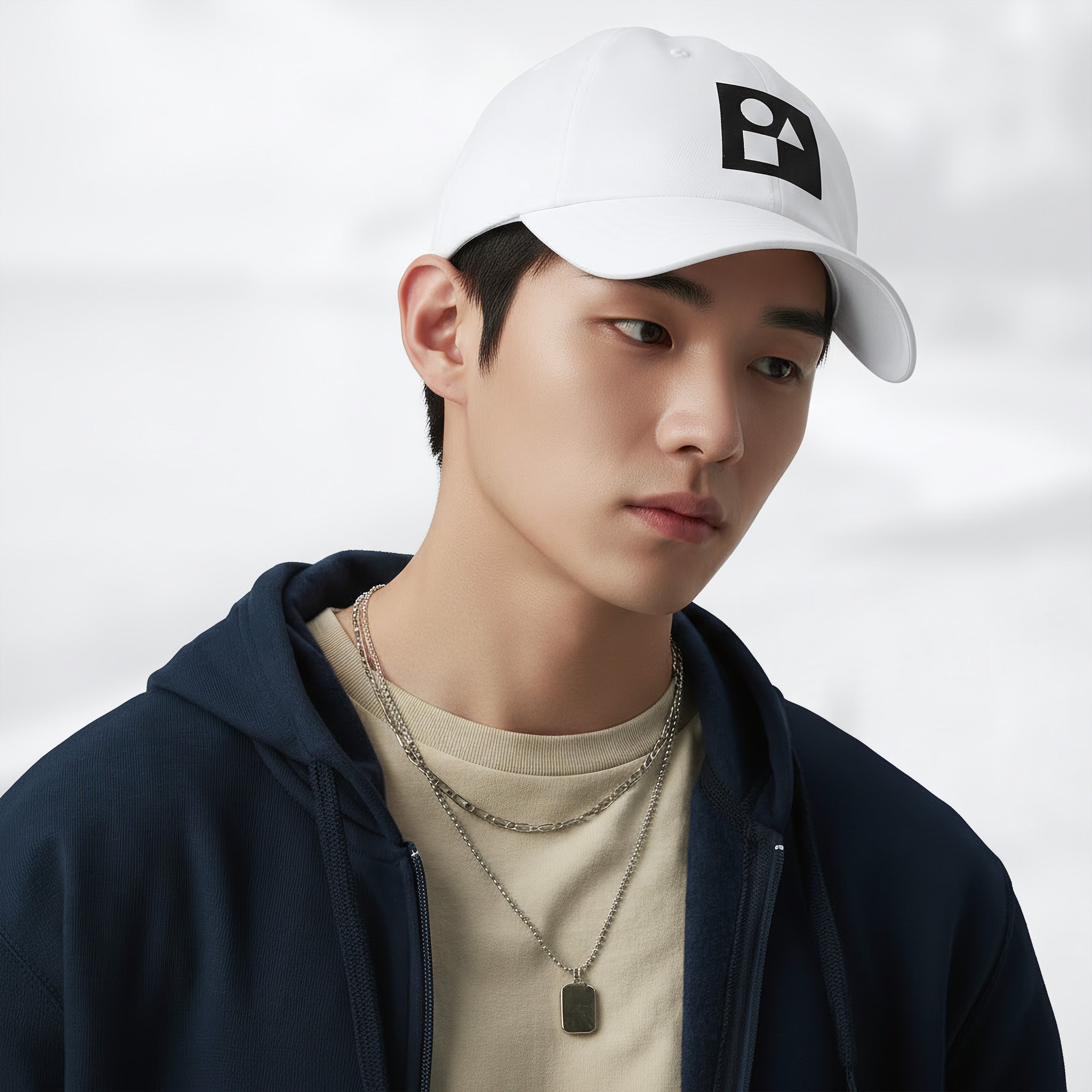 Male model wearing the white Under Armour x Form Fundamentals Stamp Cap (No.19) in a three‑quarter front pose; the cap’s curved brim and black Form Fundamentals logo are visible, styled with a navy hoodie over a beige top and layered chain in a neutral studio setting.