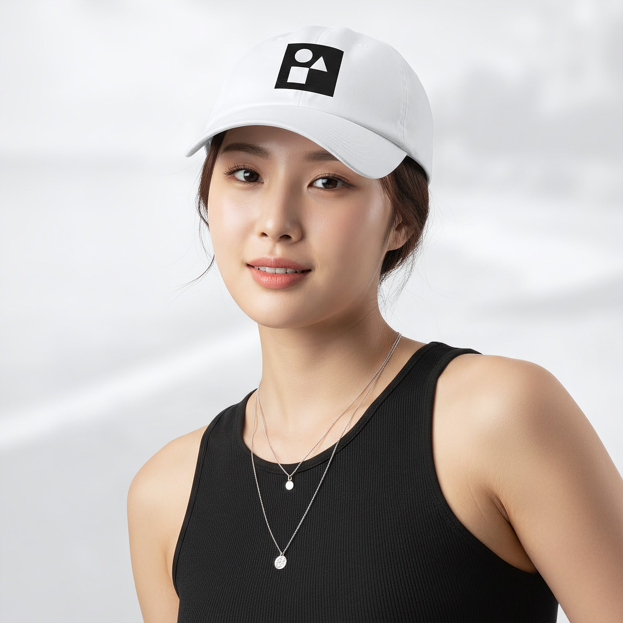 Female model wearing the white Under Armour x Form Fundamentals Stamp Cap (No.19); front view of the cap with black geometric emblem and curved brim, paired with a black tank top and layered necklaces against a softly blurred background.