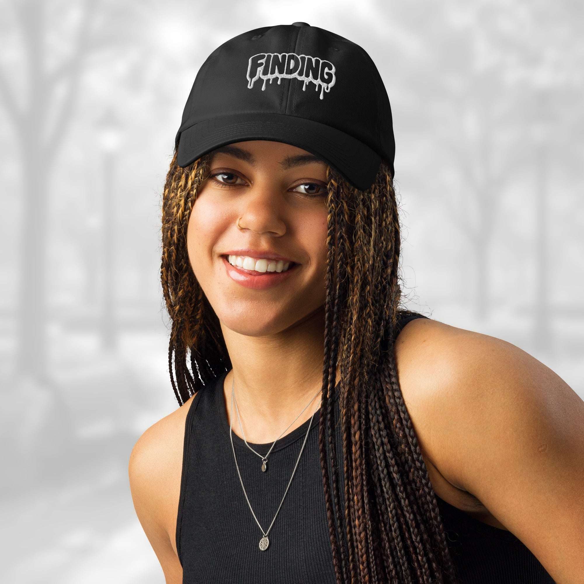 Front portrait of a smiling woman in the Under Armour x Title-3 Finding Logo Cap; the black cap’s bold white graffiti Finding embroidery sits above the curved visor, paired with layered necklaces and long braids.