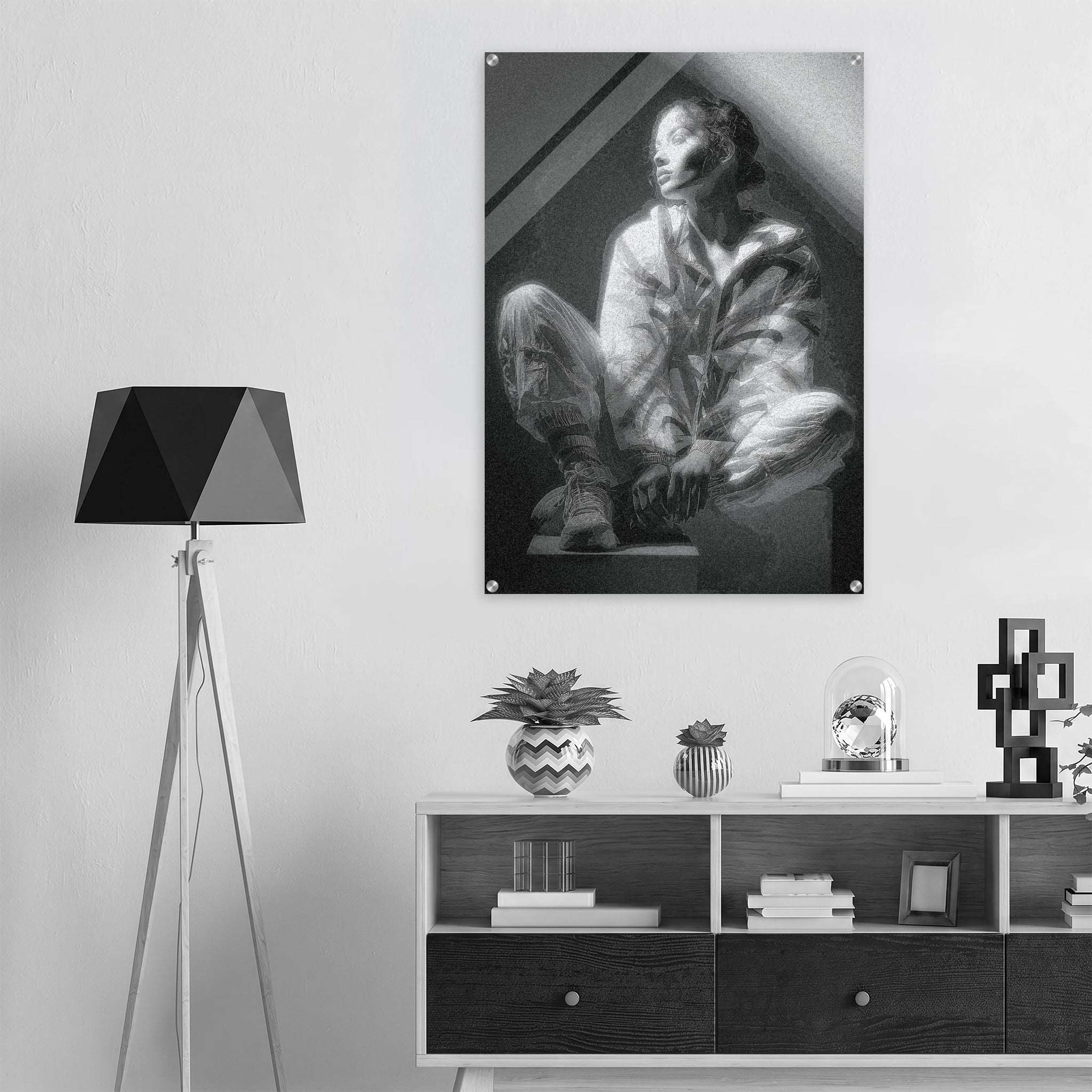 A black and white art print depicting a seated figure in a dramatic interior, displayed on a light gray wall above a modern white and dark wood cabinet, next to a black tripod floor lamp.